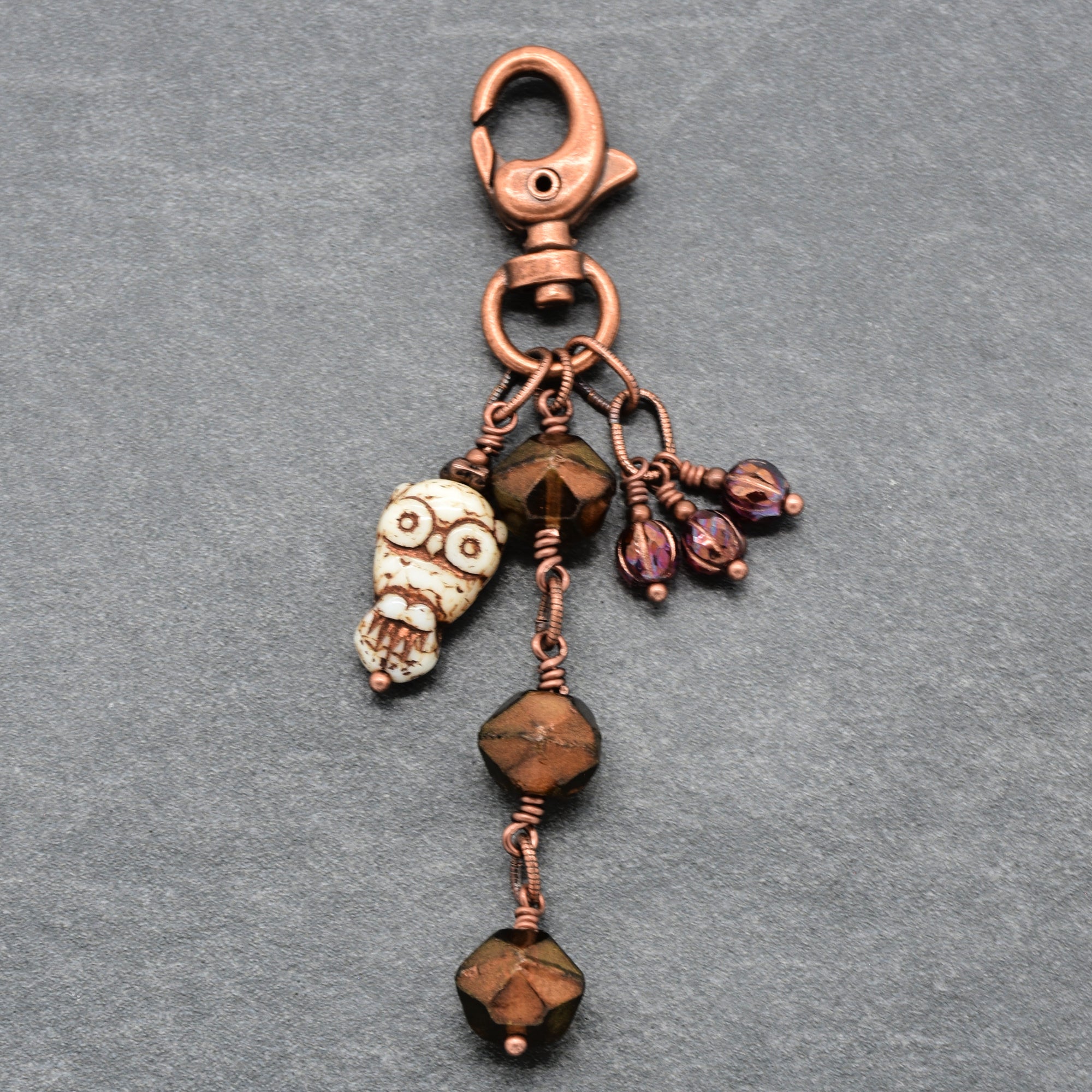 A handmade copper keychain featuring a lobster clasp, an owl charm, faceted brown beads, and small burgundy charms, displayed on a textured gray background - made with love in Colorado by Ruby Acorn.