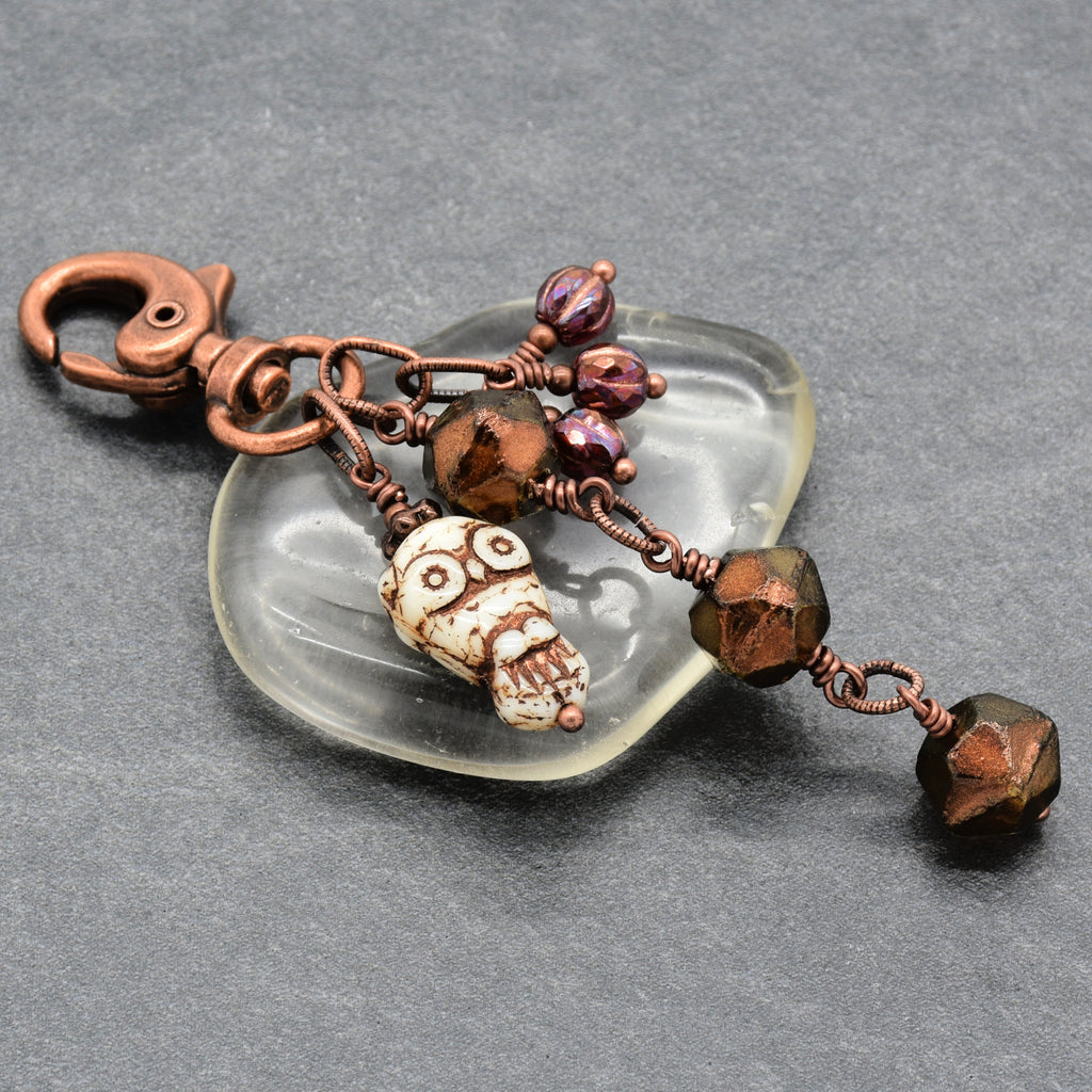A handmade copper keychain featuring a carved owl charm, Czech glass English cut faceted bronze beads, and iridescent purple beads, displayed on a translucent glass dish against a dark gray background - made with love in Colorado by Ruby Acorn.