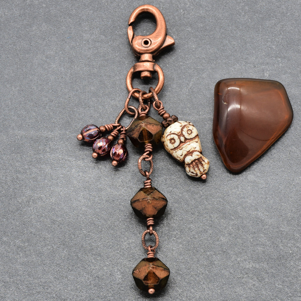 A handmade copper keychain featuring a lobster clasp, a small owl charm, faceted brown beads, and burgundy glass beads next to a polished piece of glass, displayed on a textured gray background- made with love in Colorado by Ruby Acorn.