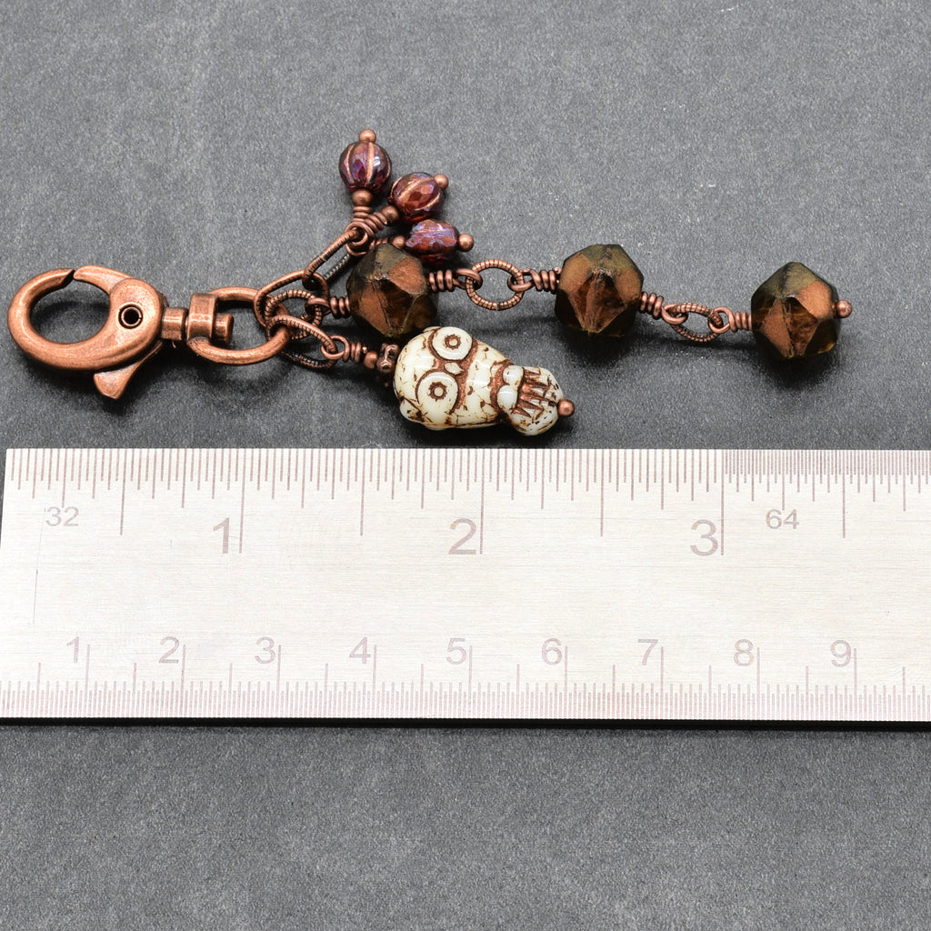A handmade copper wire keychain charm with an owl bead, faceted beads, and a lobster clasp, shown next to a metal ruler for scale on a dark gray textured background - made with love in Colorado by Ruby Acorn.