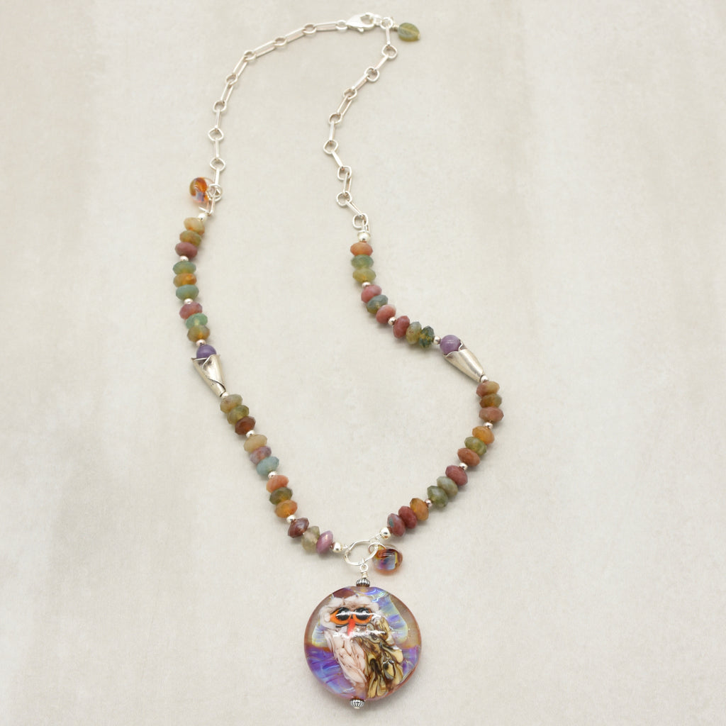 A beaded necklace with an artisan lampwork glass pendant featuring a colorful illustration of an owl set against a neutral light beige background - made. with love in Colorado by Ruby Acorn.