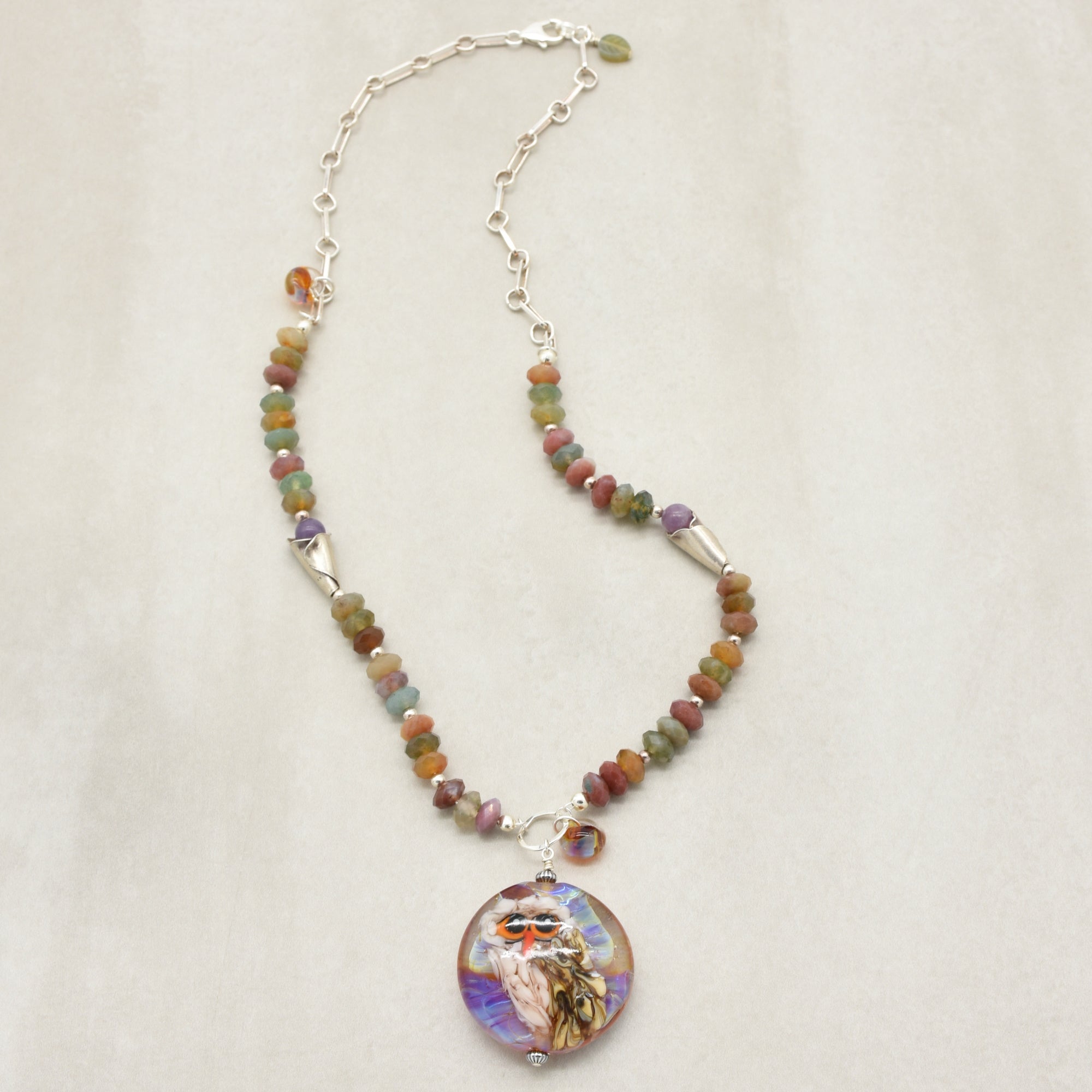 A beaded necklace with an artisan lampwork glass pendant featuring a colorful illustration of an owl set against a neutral light beige background - made. with love in Colorado by Ruby Acorn.