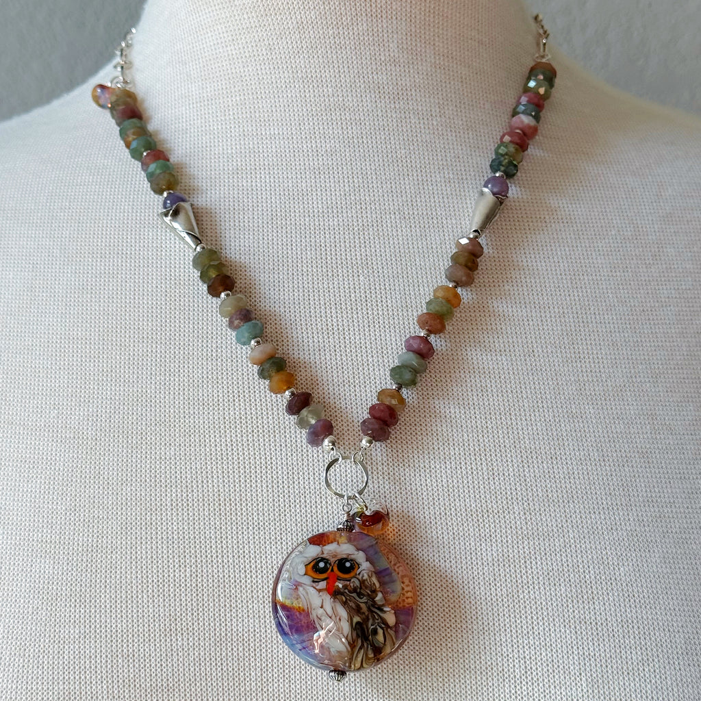 A beaded necklace displayed on a cream fabric mannequin bust. The necklace features a strand of small, multicolored beads in earthy tones, and an artisan lampwork glass pendant depicting an owl with orange eyes and a purple, white, and brown patterned background - made with love in Colorado by Ruby Acorn.