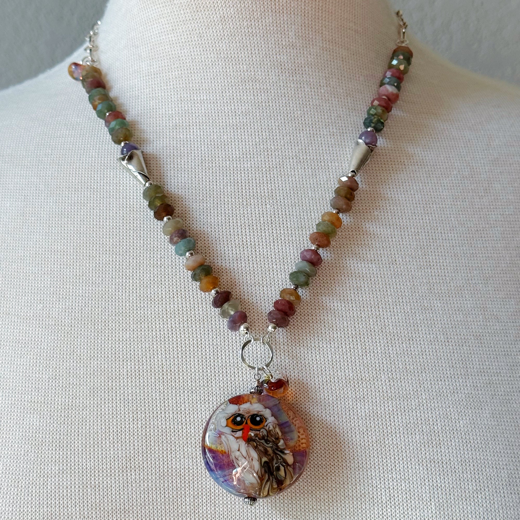A beaded necklace displayed on a cream fabric mannequin bust. The necklace features a strand of small, multicolored beads in earthy tones, and an artisan lampwork glass pendant depicting an owl with orange eyes and a purple, white, and brown patterned background - made with love in Colorado by Ruby Acorn.