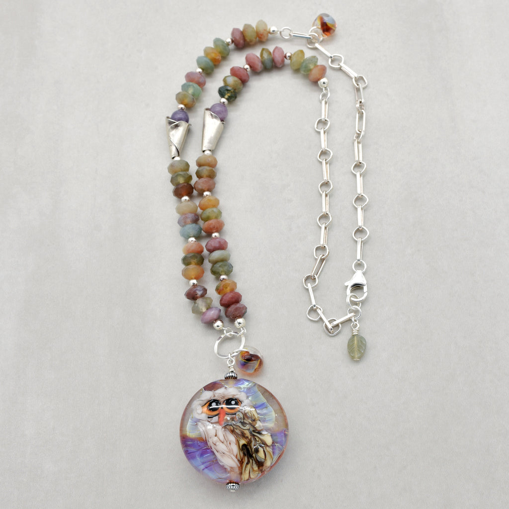 A beaded necklace with an artisan lampwork glass pendant featuring a painted owl, displayed on a light gray background. The beads are multicolored in autumn hued shades, and the necklace includes silver chain and silver lobster claw - made with love in Colorado by Ruby Acorn.