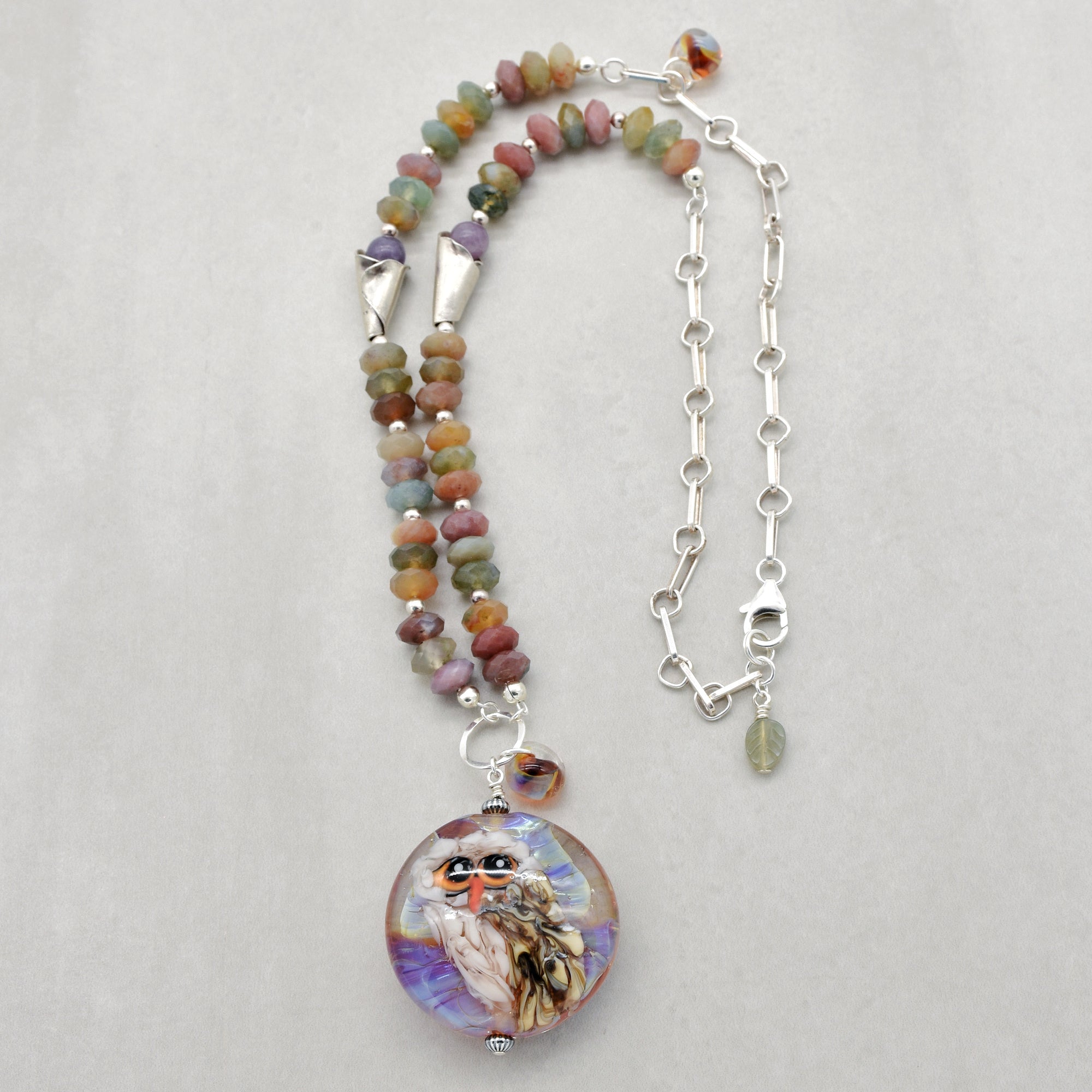 A beaded necklace with an artisan lampwork glass pendant featuring a painted owl, displayed on a light gray background. The beads are multicolored in autumn hued shades, and the necklace includes silver chain and silver lobster claw - made with love in Colorado by Ruby Acorn.