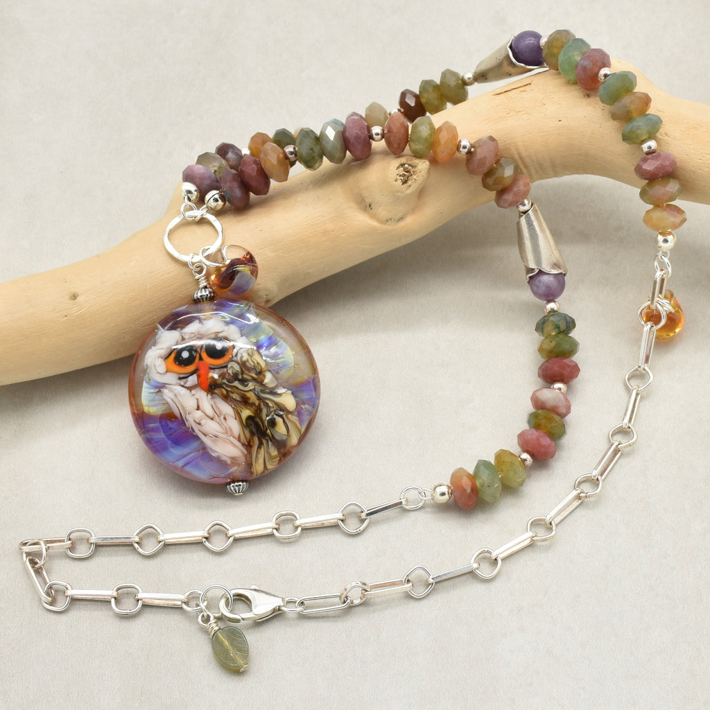 A beaded necklace with an artisan lampwork glass pendant featuring an owl illustration, displayed on a light wood branch against a neutral background - made with love in Colorado by Ruby Acorn.