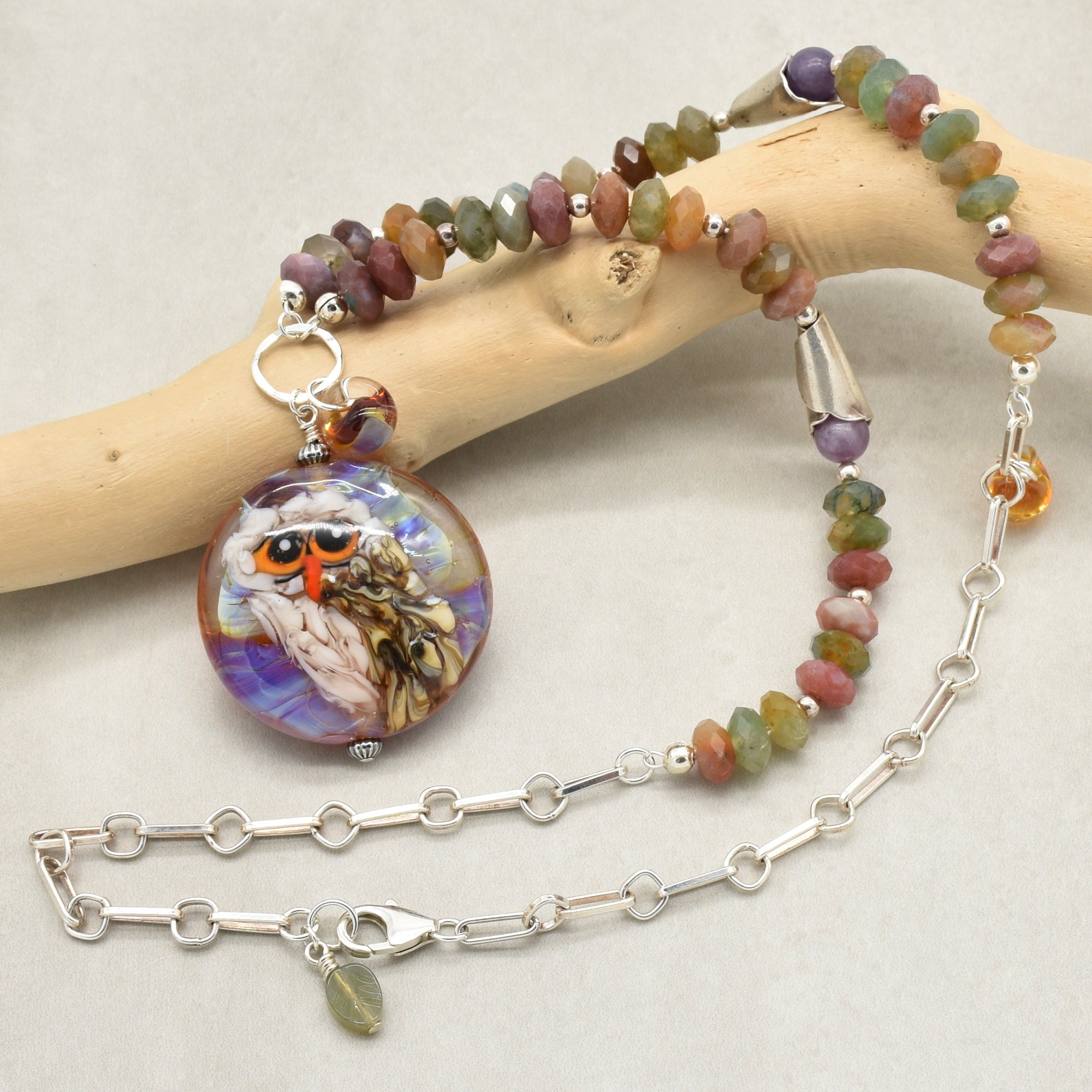 A beaded necklace with an artisan lampwork glass pendant featuring an owl illustration, displayed on a light wood branch against a neutral background - made with love in Colorado by Ruby Acorn.