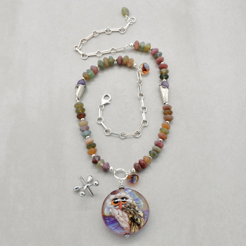 A beaded necklace with an artisan lampwork glass pendant featuring an owl, displayed on a light gray background. The necklace features multi-colored natural stone beads, silver-toned chain, and a silver lobster clasp - made with love in Colorado by Ruby Acorn.