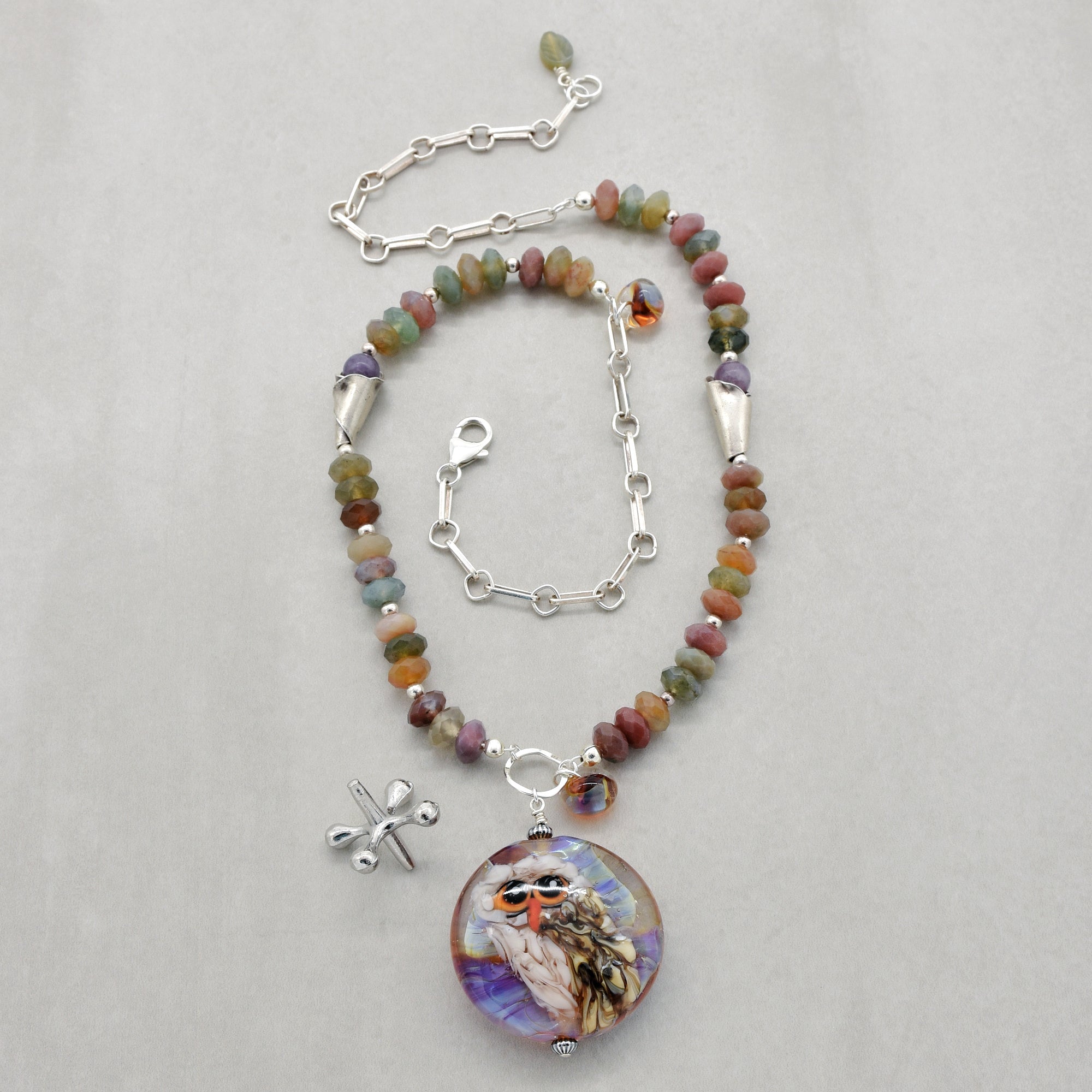 A beaded necklace with an artisan lampwork glass pendant featuring an owl, displayed on a light gray background. The necklace features multi-colored natural stone beads, silver-toned chain, and a silver lobster clasp - made with love in Colorado by Ruby Acorn.
