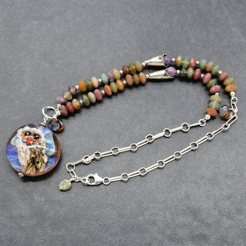 A beaded necklace with a round owl illustration pendant, featuring multicolored Indian agate beads, silver chain, and a silver lobster claw on a dark gray textured background - made with love in Colorado by Ruby Acorn.