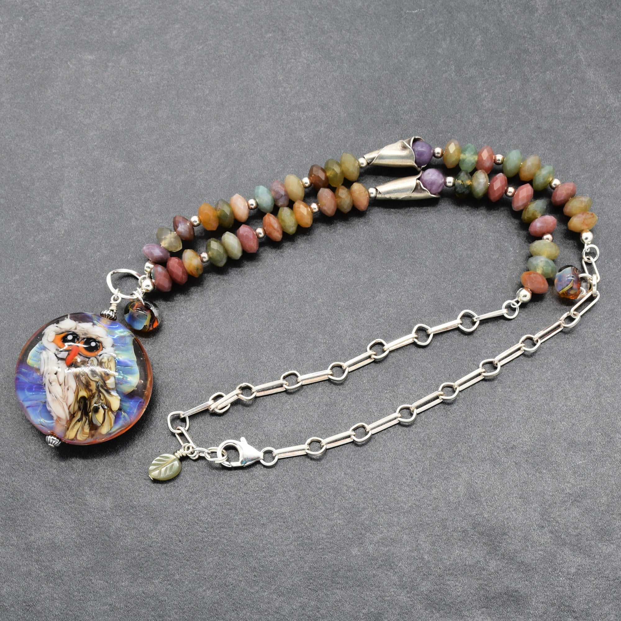 A beaded necklace with a round owl illustration pendant, featuring multicolored Indian agate beads, silver chain, and a silver lobster claw on a dark gray textured background - made with love in Colorado by Ruby Acorn.