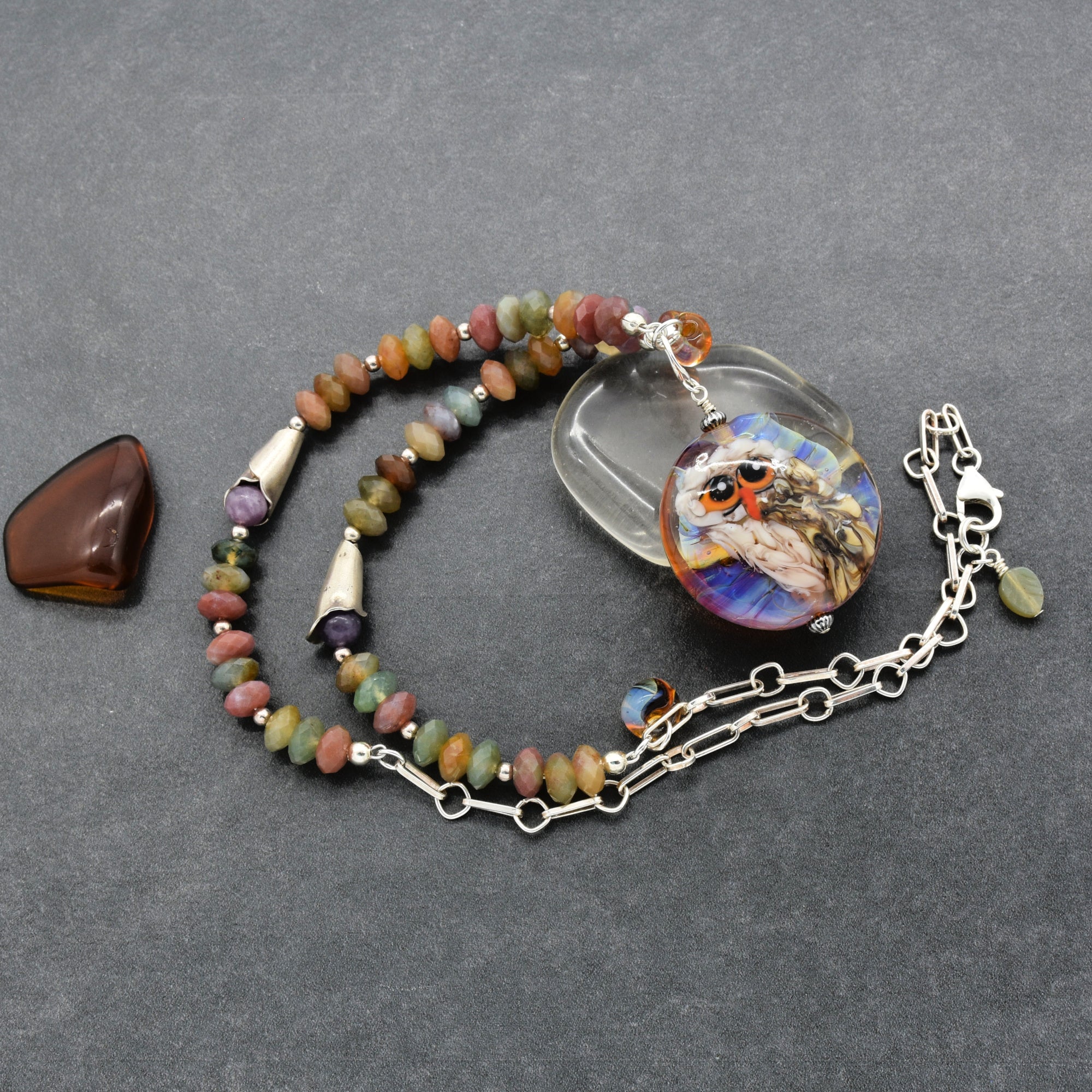 A beaded necklace with a round owl pendant, displayed on a dark gray textured background next to a polished brown stone. The necklace features multi-colored beads in shades of green, brown, orange, and purple, silver spacers, and a silver chain with a lobster clasp - made with love in Colorado by Ruby Acorn.