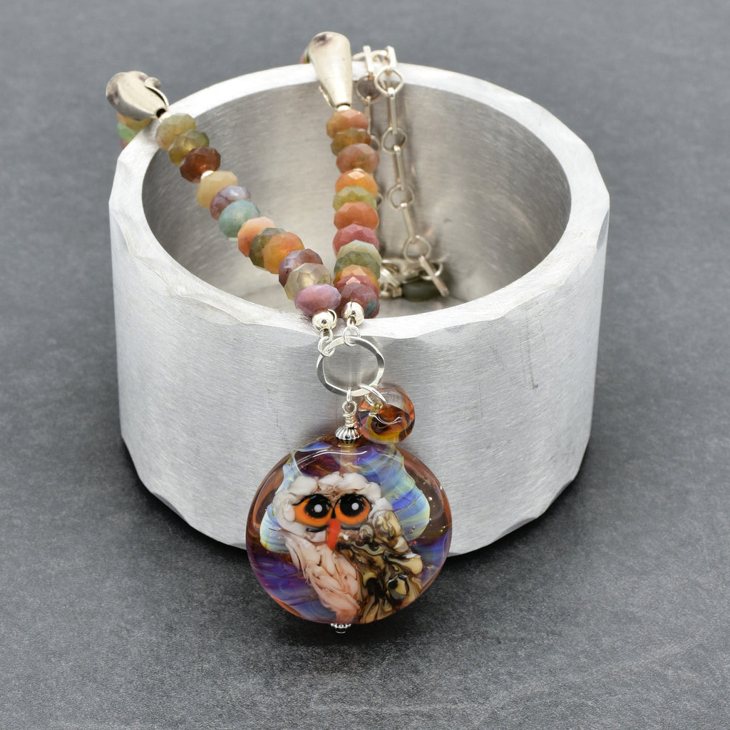 A beaded necklace with a colorful owl pendant, displayed on a textured silver cup against a dark gray background - made with love in Colorado by Ruby Acorn.
