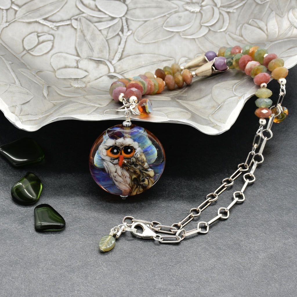 A beaded necklace with an artisan lampwork glass pendant depicting an owl, displayed on a textured silver tray on a dark gray background - made with love in Colorado by Ruby Acorn.