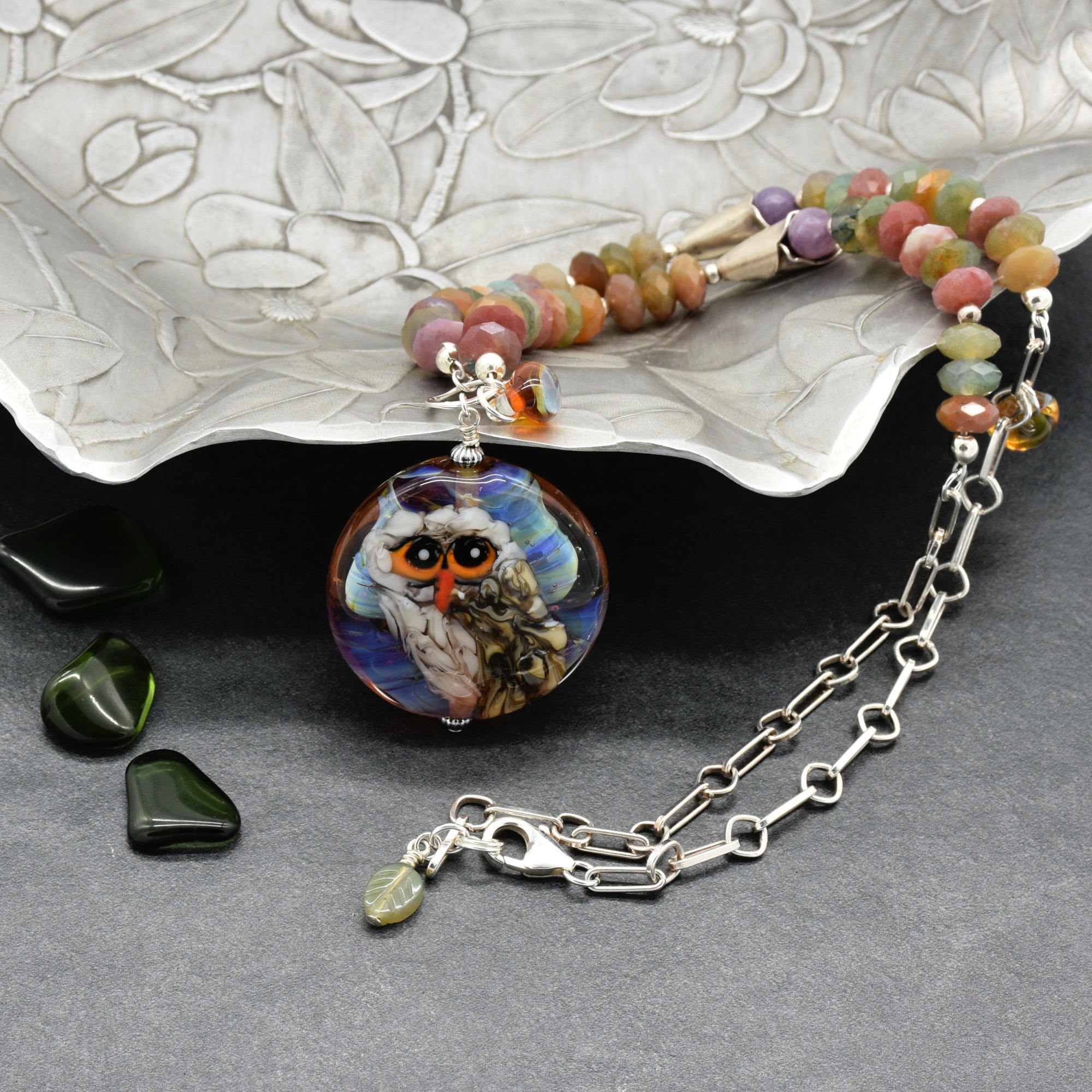 A beaded necklace with an artisan lampwork glass pendant depicting an owl, displayed on a textured silver tray on a dark gray background - made with love in Colorado by Ruby Acorn.