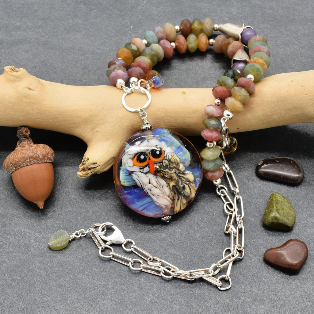A beaded necklace with an artisan lampwork glass owl pendant, displayed on a light wood branch over a dark gray background. The necklace features multicolored earthy hued Indian agate beads, silver chain, and a silver lobster claw - made with love in Colorado by Ruby Acorn.