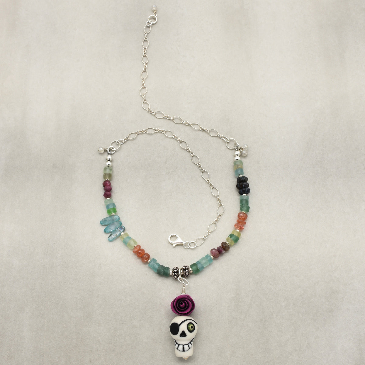 A beaded necklace with a sugar skull pendant, featuring a mix of colorful translucent beads in blue, green, orange, and purple, accented by silver-toned spacers and a purple rose charm, displayed on a neutral textured background - made with love in Colorado by Ruby Acorn.