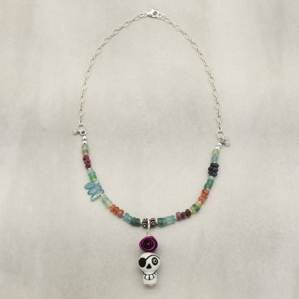 A beaded necklace with a sugar skull pendant, featuring a silver chain, multicolor faceted beads, a purple paper rose charm, and a white sugar skull pendant with black and green details, displayed on a neutral textured background - made with love in Colorado by Ruby Acorn.