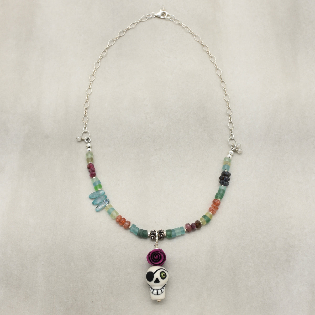 A beaded necklace with a sugar skull pendant, featuring a silver chain, multicolor faceted beads, a purple paper rose charm, and a white sugar skull pendant with black and green details, displayed on a neutral textured background - made with love in Colorado by Ruby Acorn.