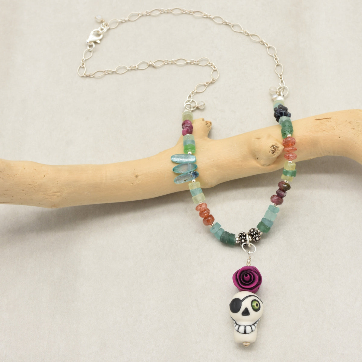 A beaded necklace with a sugar skull pendant, displayed on a light wood branch against a neutral light gray background. The necklace features a mix of colorful beads in green, blue, orange, purple, and silver-toned metal chain and charms - made with love in Colorado by Ruby Acorn.