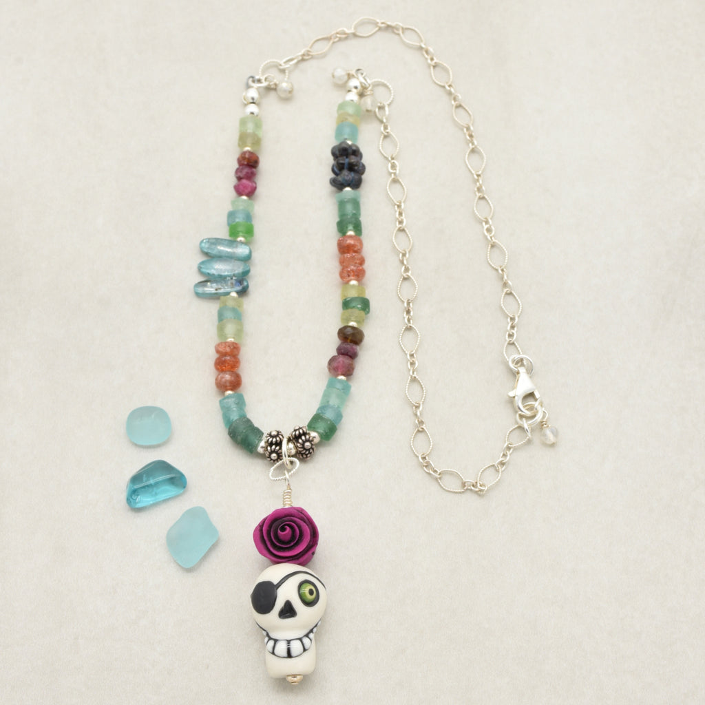 A handmade beaded necklace with a sugar skull pendant, displayed on a light beige textured background. The necklace features a mix of colorful glass beads (green, blue, orange, purple, red) and silver chain links, with three small light blue glass chips placed to the left - made with love in Colorado by Ruby Acorn.