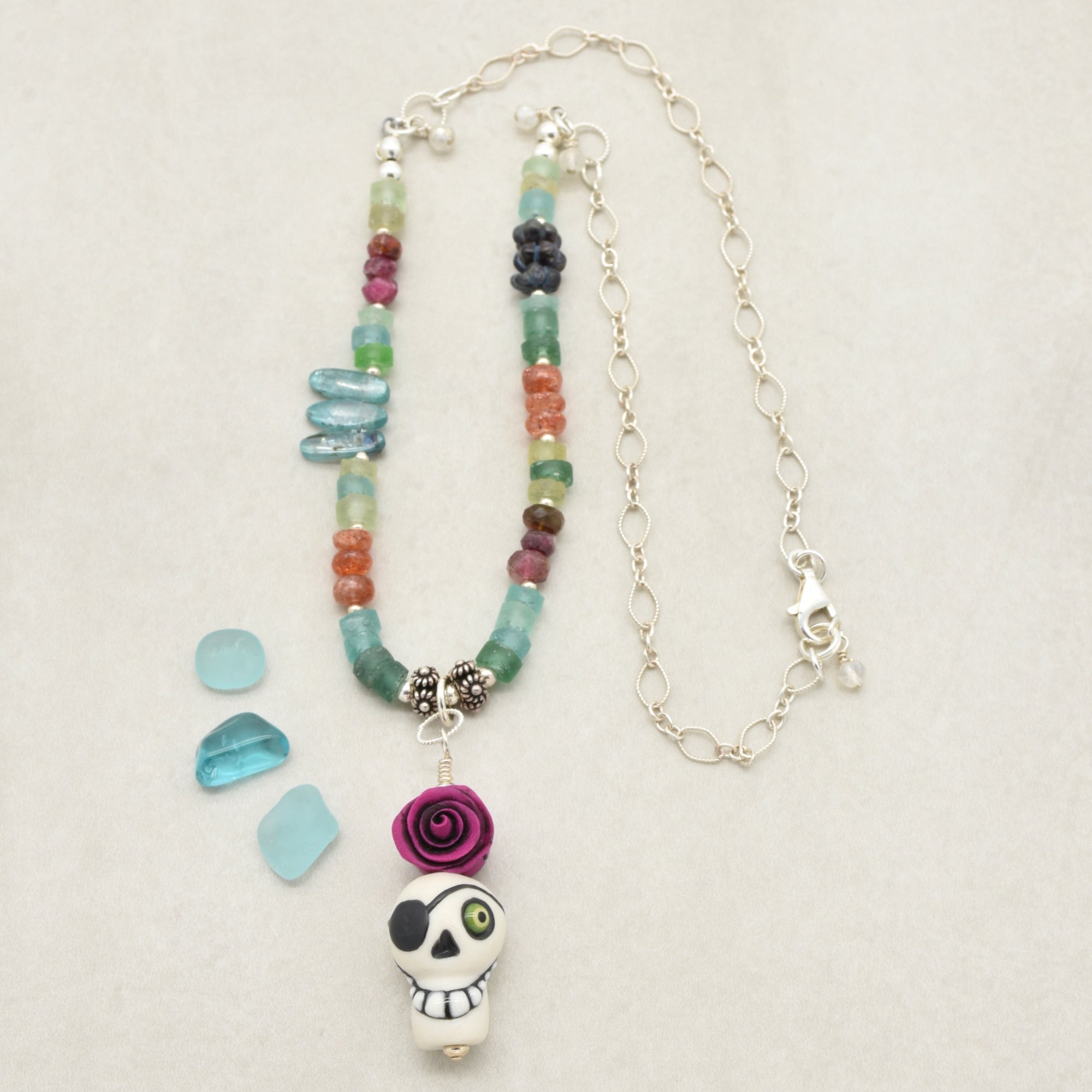 A handmade beaded necklace with a sugar skull pendant, displayed on a light beige textured background. The necklace features a mix of colorful glass beads (green, blue, orange, purple, red) and silver chain links, with three small light blue glass chips placed to the left - made with love in Colorado by Ruby Acorn.