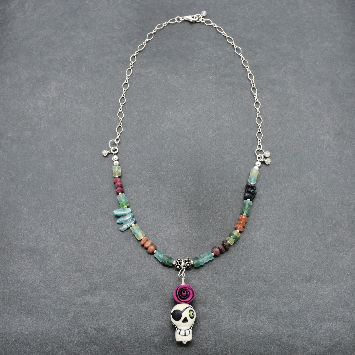 A beaded necklace with a silver chain, featuring colorful stone beads, a purple rolled rose charm, and a white skull pendant, displayed on a dark gray textured background - made with love in Colorado by Ruby Acorn.