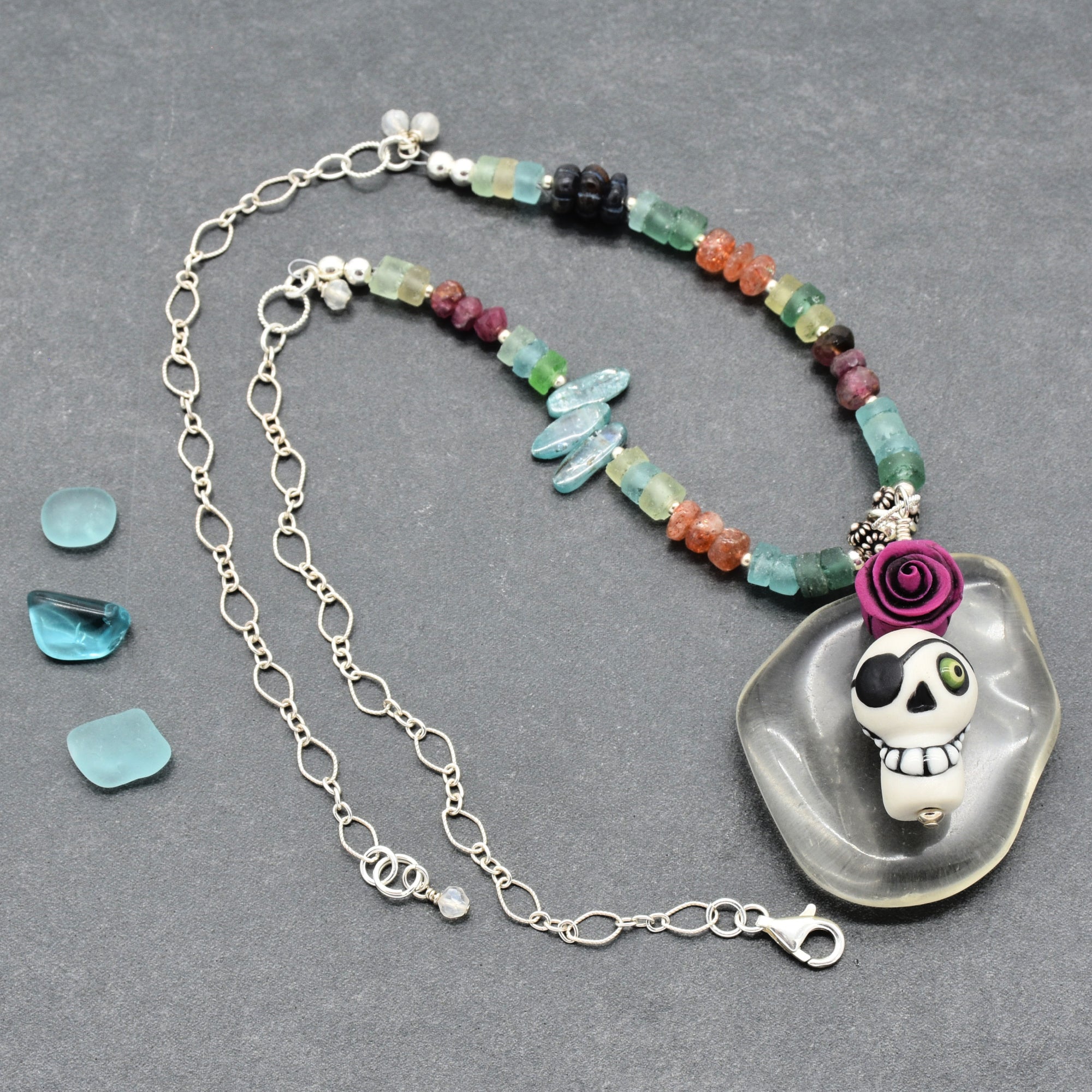 A beaded necklace with a skull pendant and silver chain, displayed on a dark gray textured background. The necklace features a variety of colorful faceted beads, a silver chain, a clear resin pendant, and a purple rose accent - made with love in Colorado by Ruby Acorn.