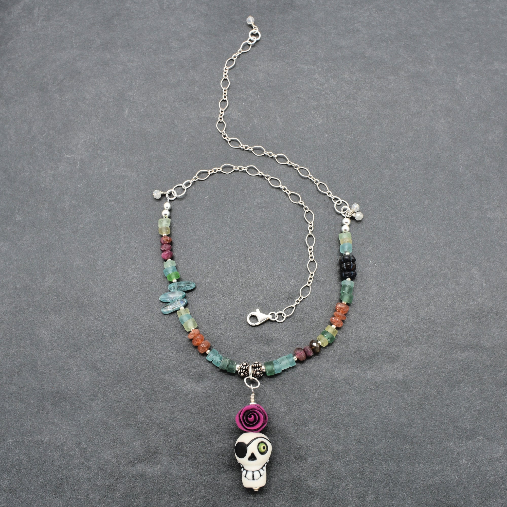 A beaded necklace with a sugar skull pendant, featuring a mix of colorful beads, small flower charms, and a silver chain extender on a dark gray textured background - made with love in Colorado by Ruby Acorn.
