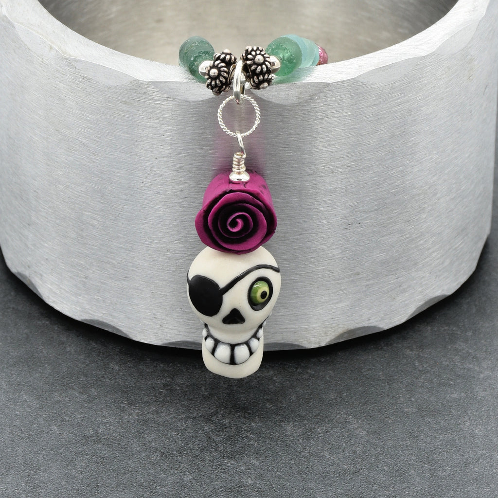 A close-up of a beaded necklace featuring a white skull charm with a black eye patch and green eye, topped with a purple rose, suspended from silver-toned beads and turquoise stones against a textured white background - made with love in Colorado by Ruby Acorn.