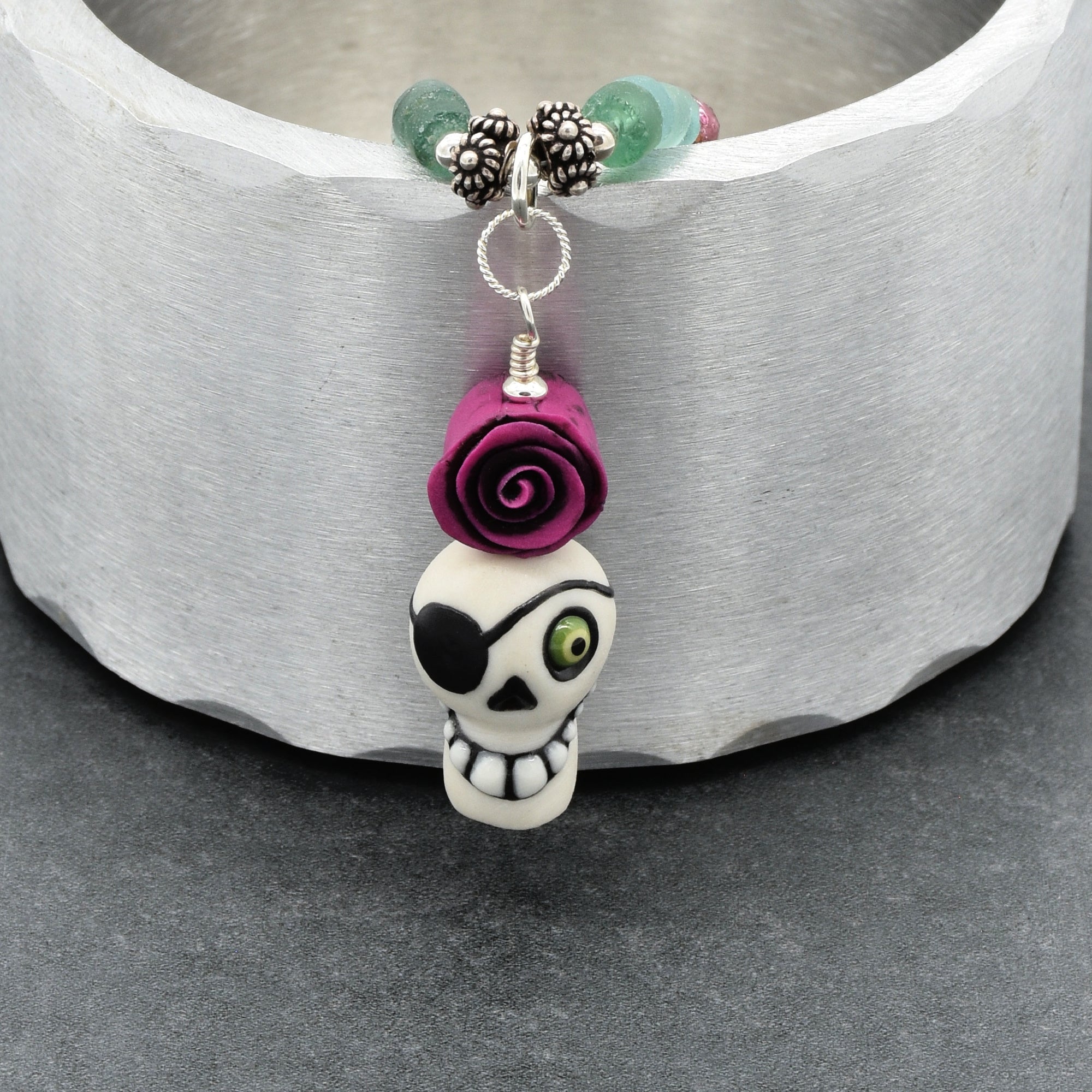 A close-up of a beaded necklace featuring a white skull charm with a black eye patch and green eye, topped with a purple rose, suspended from silver-toned beads and turquoise stones against a textured white background - made with love in Colorado by Ruby Acorn.