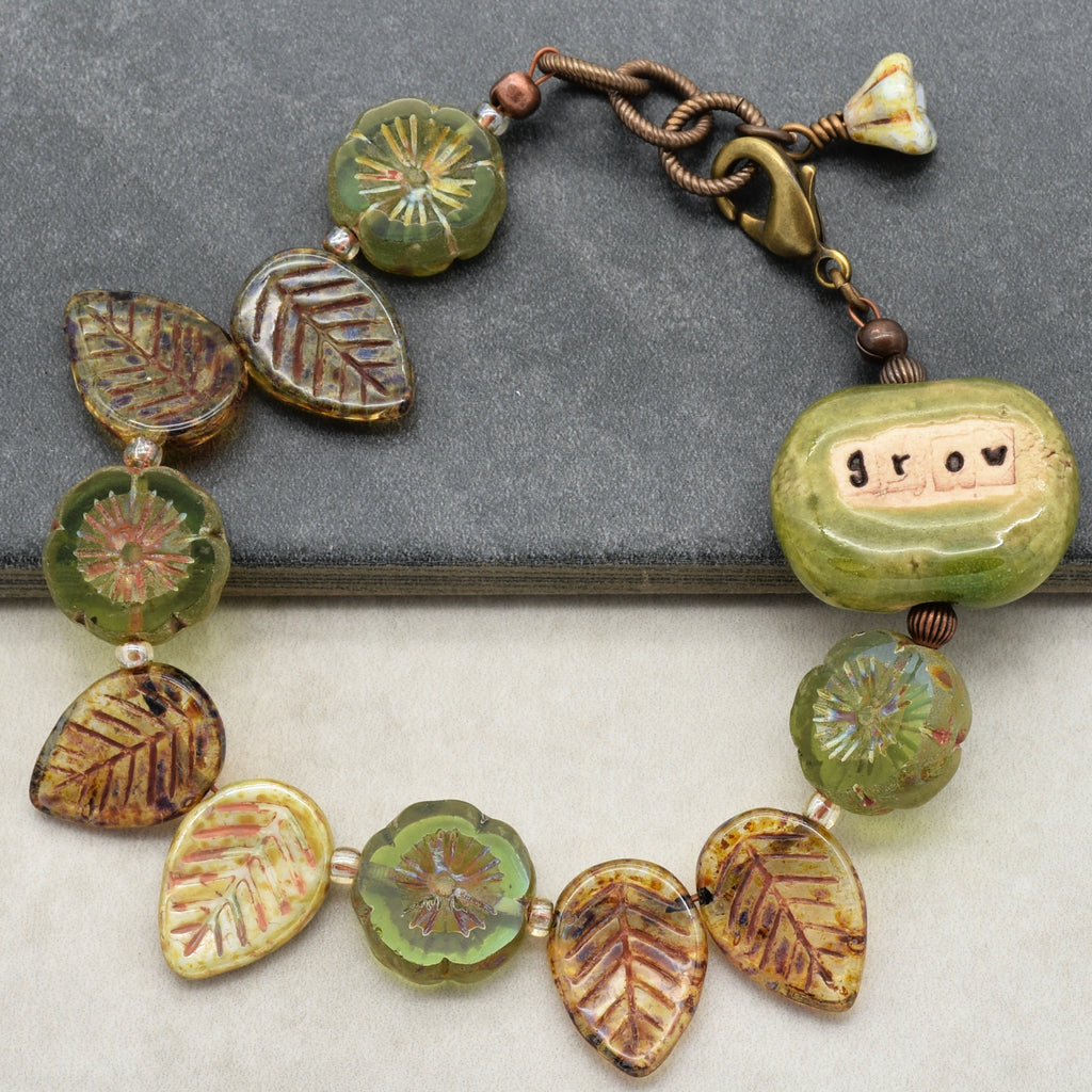 This image shows a bracelet. The size and shape of the jewelry, along with the clasp, suggest it is designed to be worn around the wrist rather than as a necklace or earring. The beads have floral and leaf designs, and there is a green bead with the word "grow" on it.
