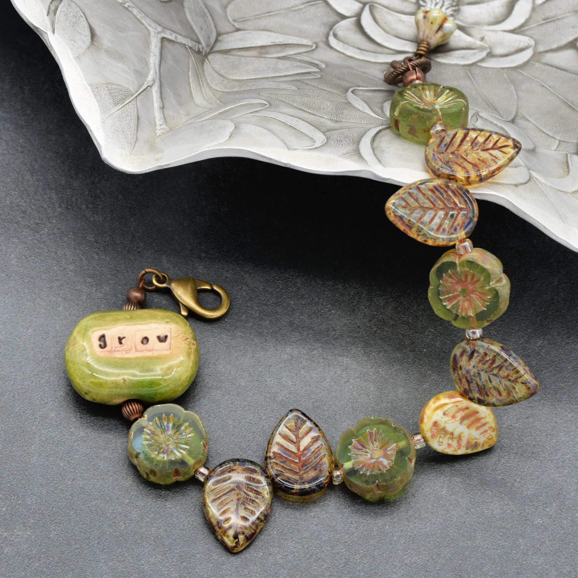 This image shows a bracelet. The jewelry has a clasp and is composed of decorative beads in the shape of leaves and flowers, with one bead featuring the word "grow" on it.