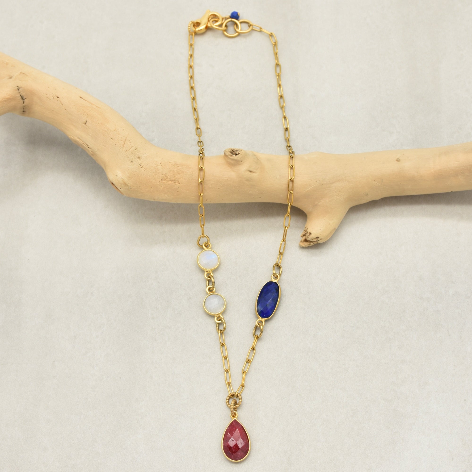Delicate gold chain necklace with a ruby pendant and lapis and rainbow moonstone links displayed on a branch - art you can wear by Ruby Acorn