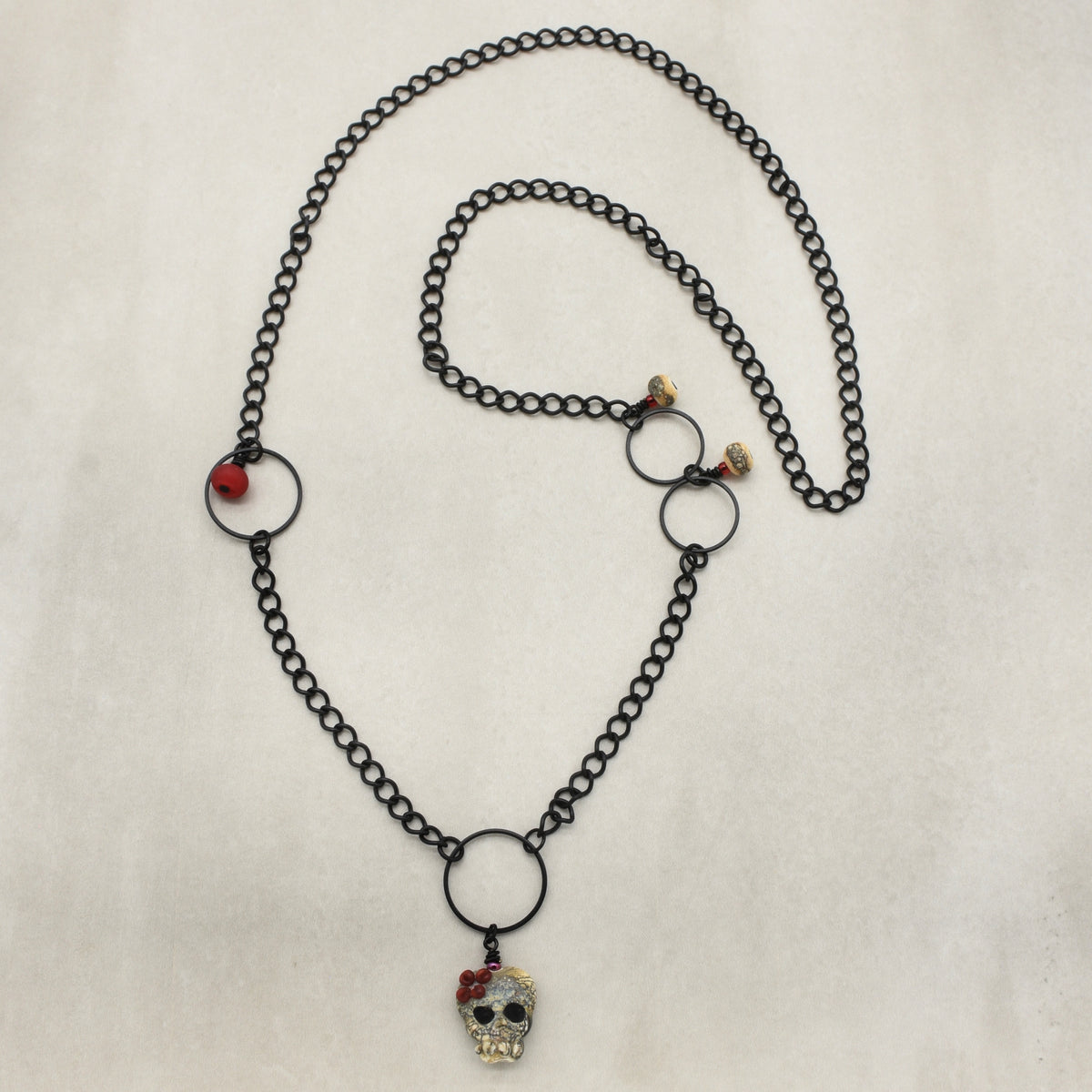 A gothic-style black chain necklace featuring a skull pendant adorned with a red flower, displayed on a textured off-white background - made with love in Colorado by Ruby Acorn.