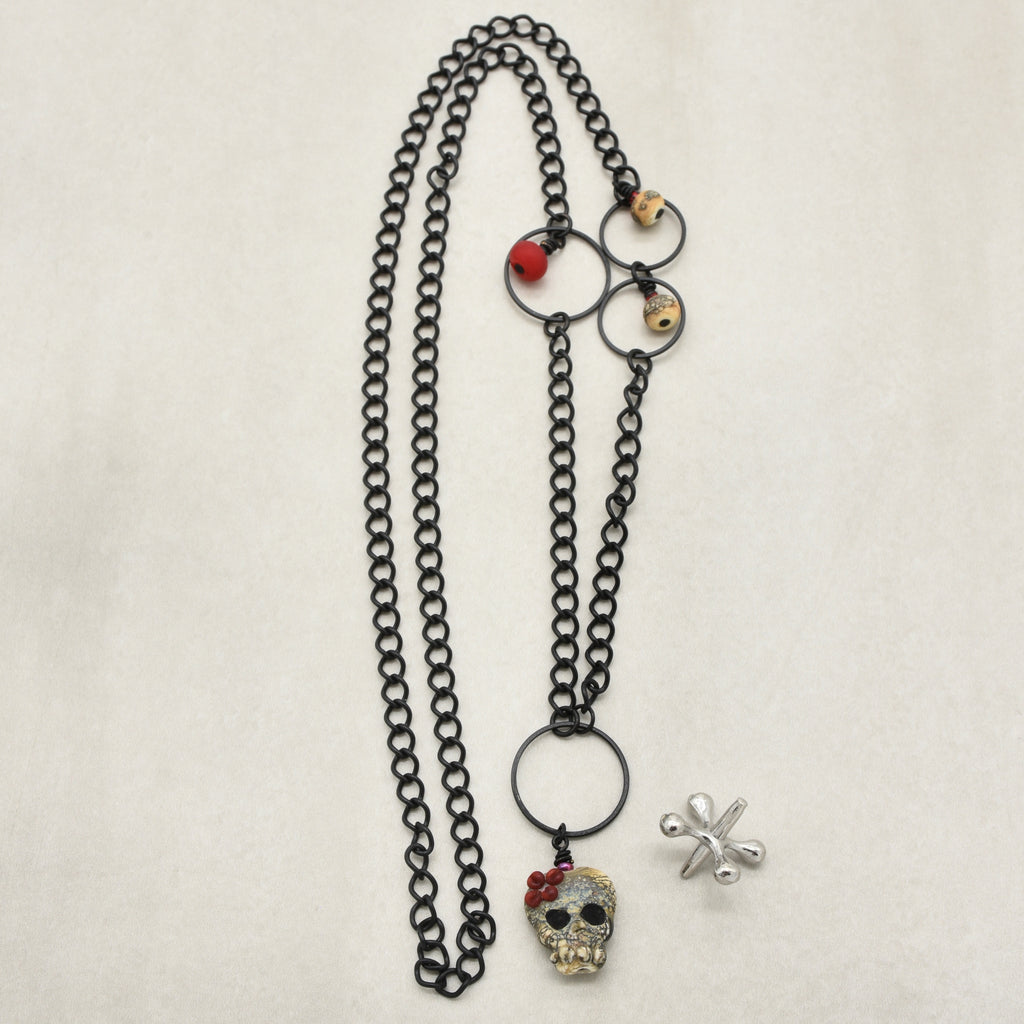 A gothic-style necklace featuring a black metal chain, a skull pendant, a silver cross charm, and small skull beads on a light beige background - made with love in Colorado by Ruby Acorn.