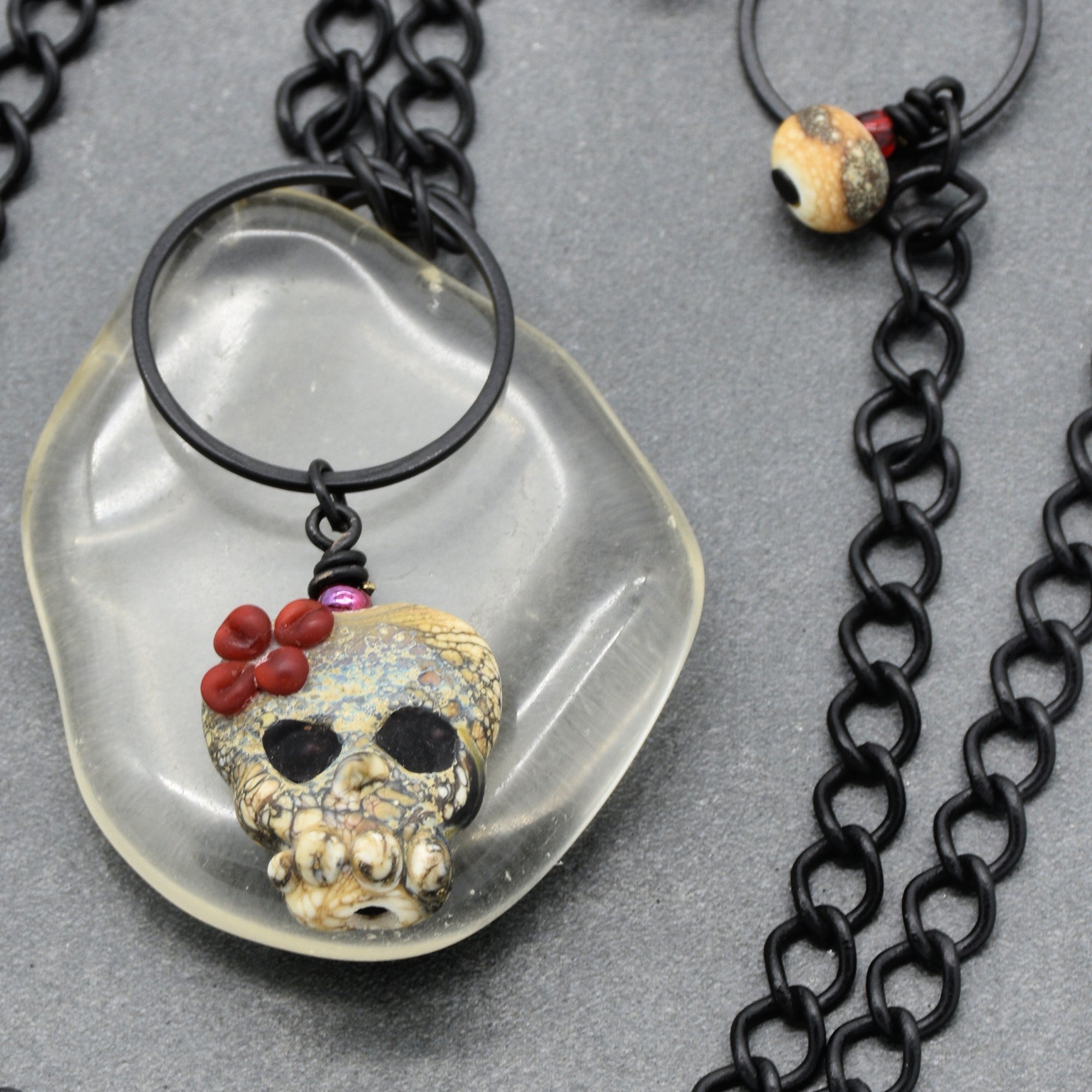A gothic-style necklace featuring a skull pendant with red flower accents, suspended in a clear glass disc over a black chain, displayed on a textured gray background - made with love in Colorado by Ruby Acorn.