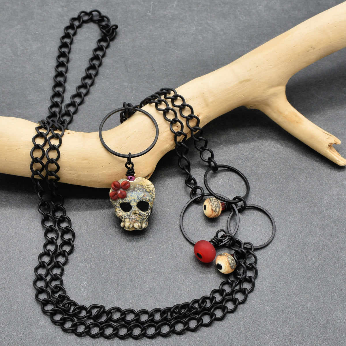 A black chain necklace with a skull pendant, red beads, and small patterned beads, displayed on a light wood branch against a gray background - made with love in Colorado by Ruby Acorn.