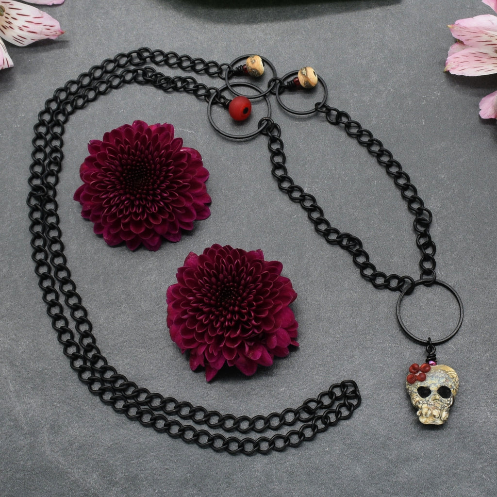 A gothic-style black chain necklace featuring a skull charm with a red flower, displayed alongside two large burgundy dahlia flowers on a textured gray background - made with love in Colorado by Ruby Acorn.