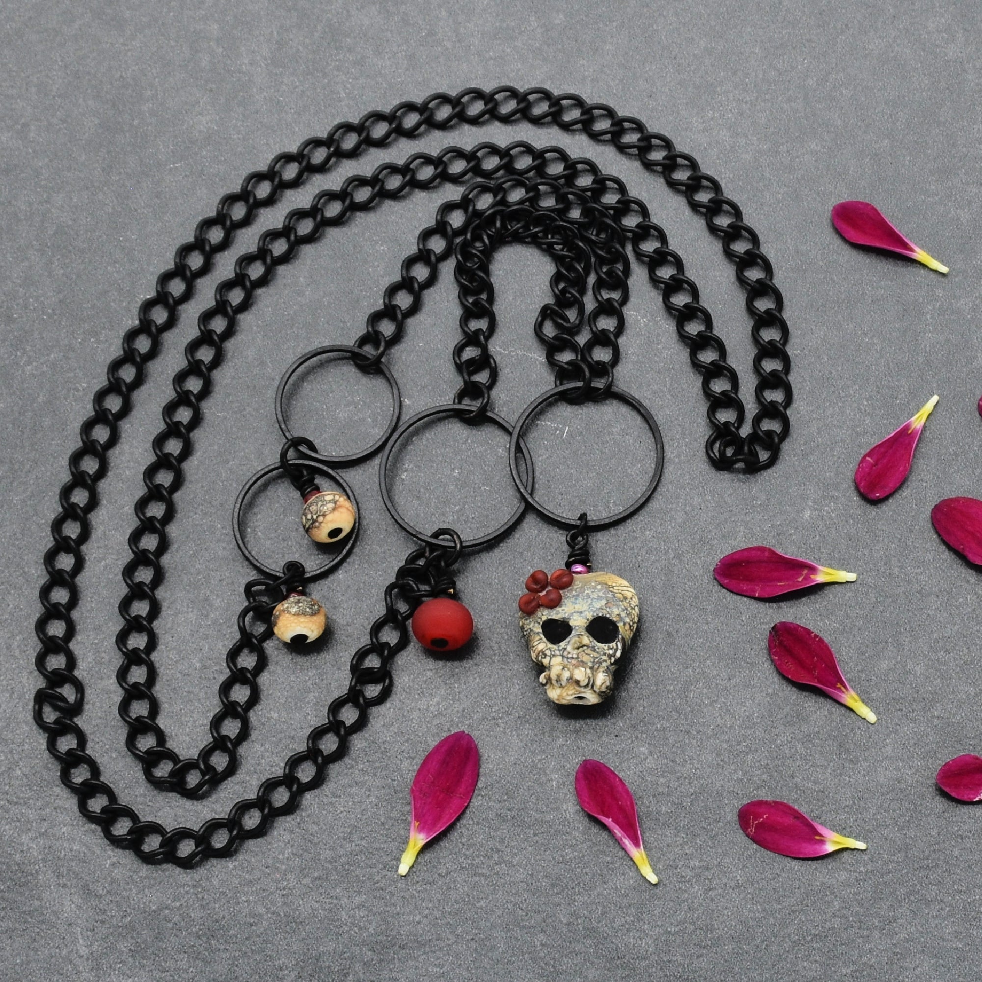 A black chain necklace laid flat on a gray textured surface, featuring three black metal rings with hanging charms: a skull pendant with red flowers, a small tan-and-black bead, and a red round bead. Pink flower petals are scattered around the necklace - made with love in Colorado by Ruby Acorn.