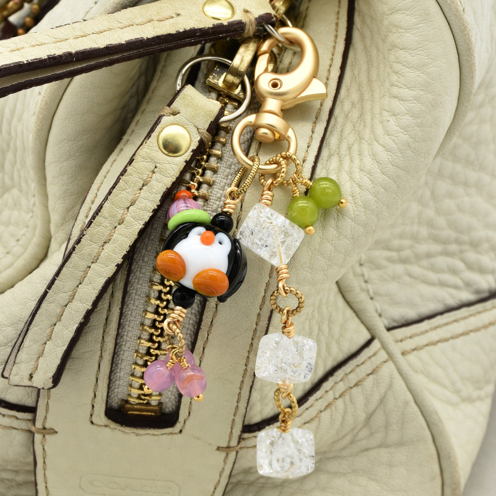 Close-up of a handbag zipper pull featuring a penguin charm with a green hat, clear faceted beads, and pink beads on a gold-tone clasp, attached to a light beige textured leather bag.
