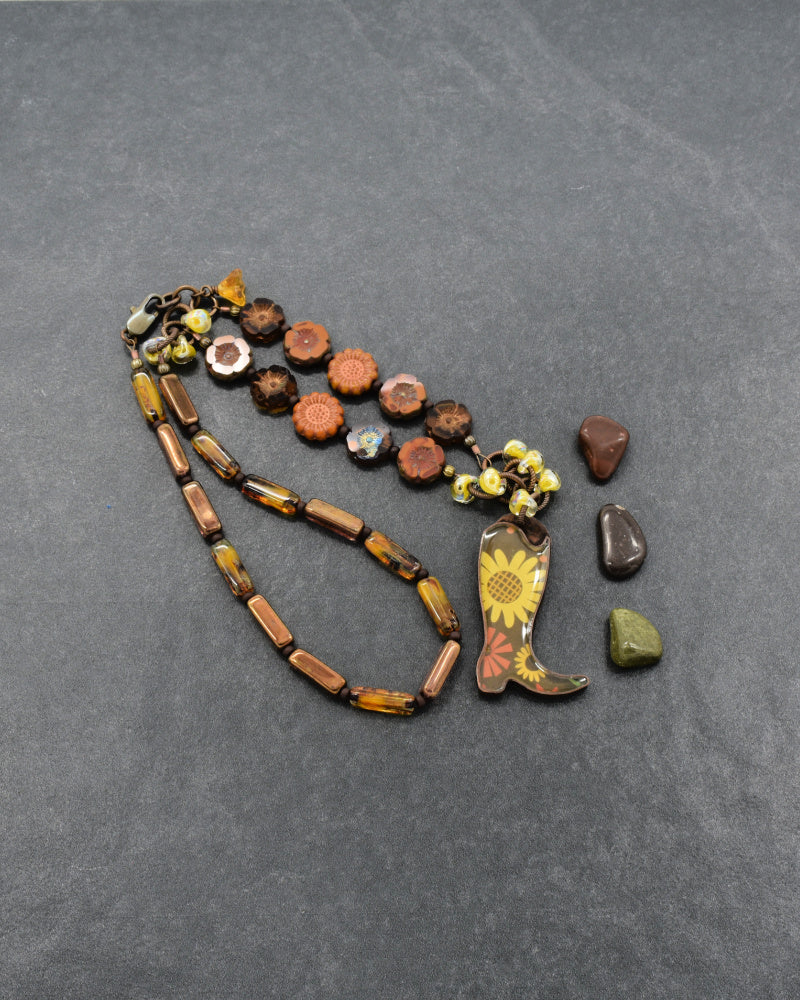 A handmade beaded necklace with a cowboy boot charm, featuring brown and amber glass beads, orange glass beads, and a yellow sunflower design on the boot, displayed on a dark gray textured background - made with love in Colorado by Ruby Acorn.