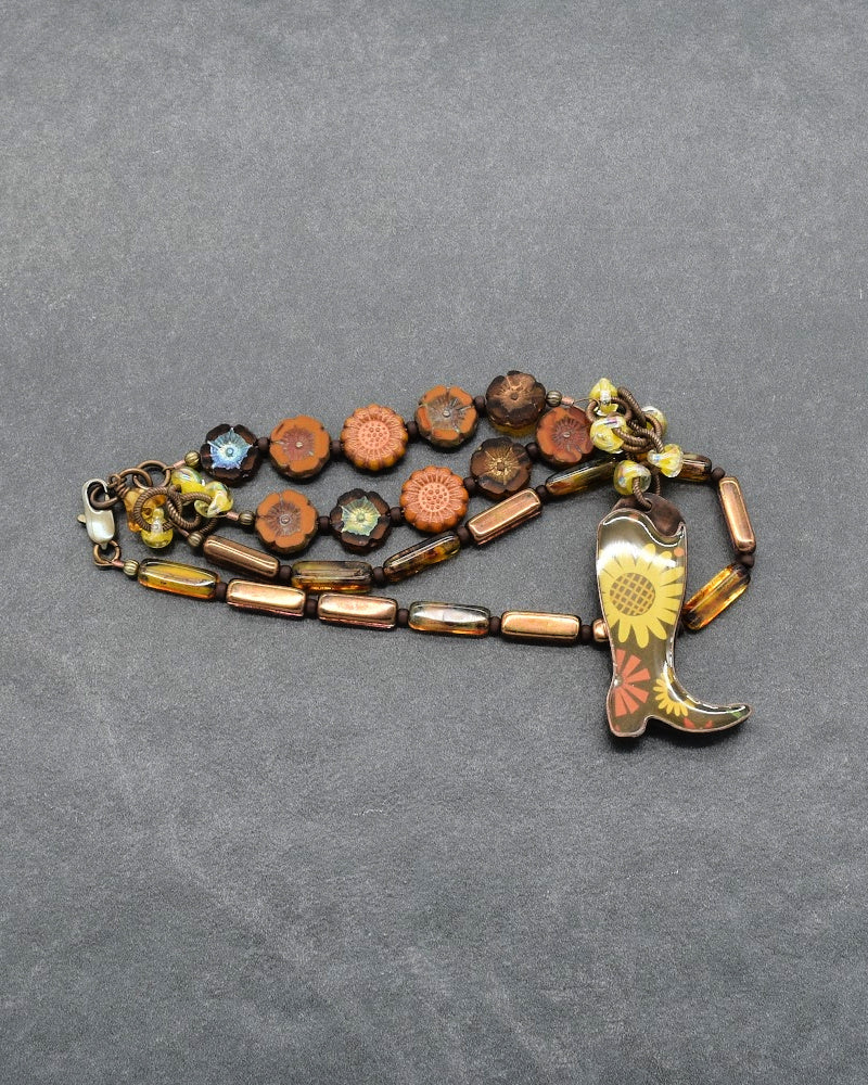 A vintage-style beaded necklace with a cowboy boot pendant, featuring a mix of brown, orange, and gold beads on a dark gray textured background.