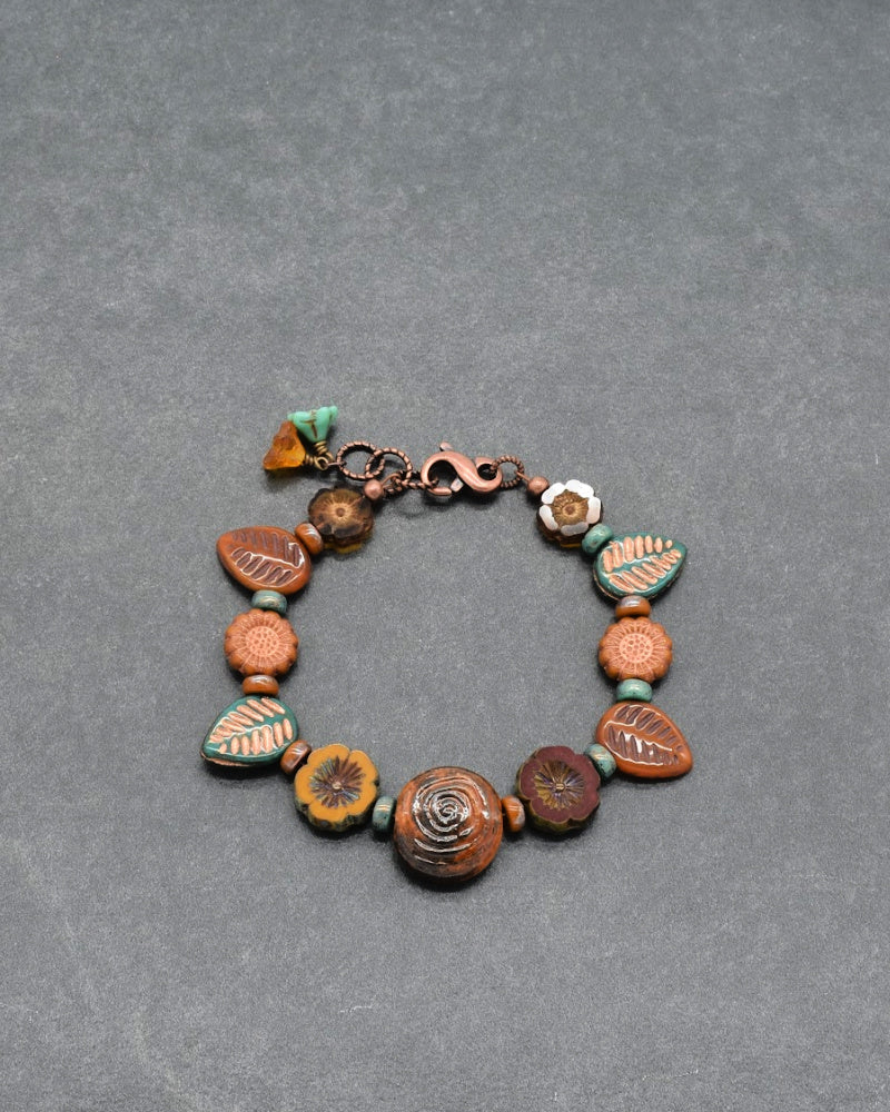 A handmade beaded bracelet featuring a variety of earthy brown, terracotta orange, and turquoise green beads with carved floral and geometric patterns, displayed on a textured gray background.