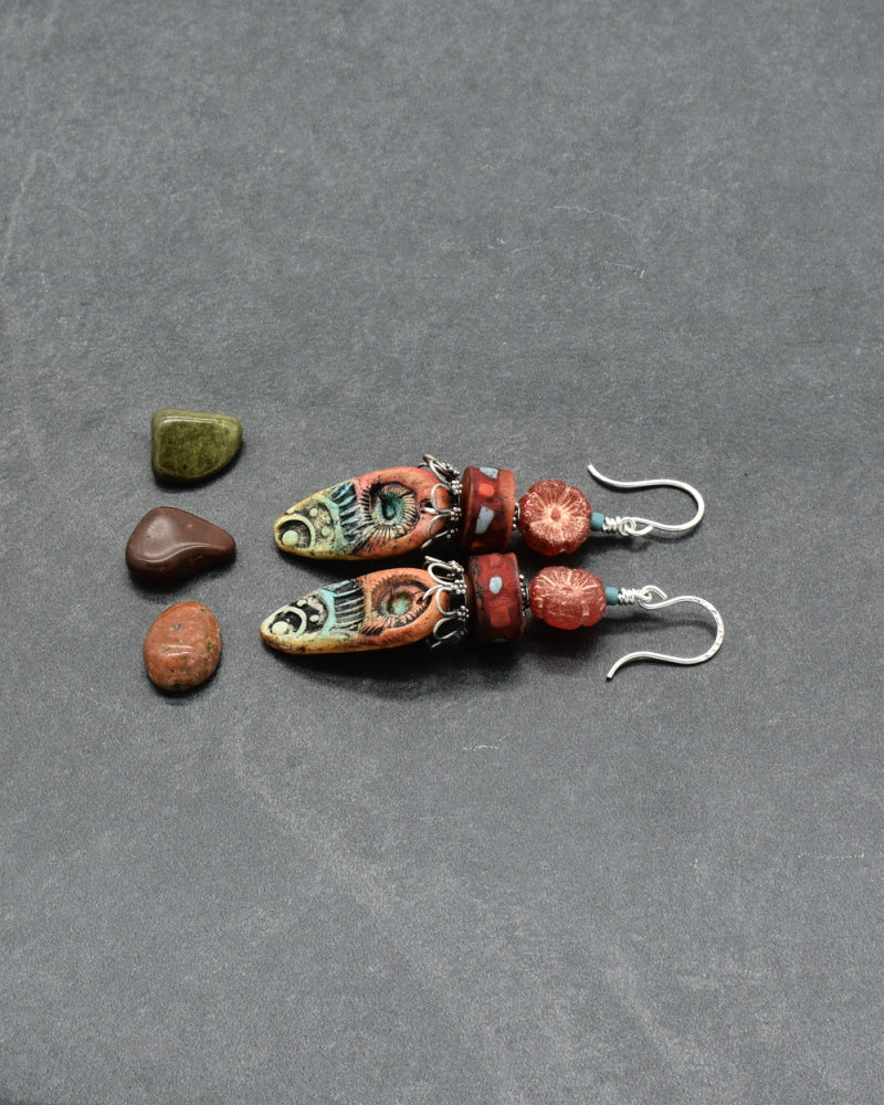 Colorful earrings with brick red and turquoise artisan charms, Bali silver beads caps and silver ear wires on a gray background - made with love by Ruby Acorn.