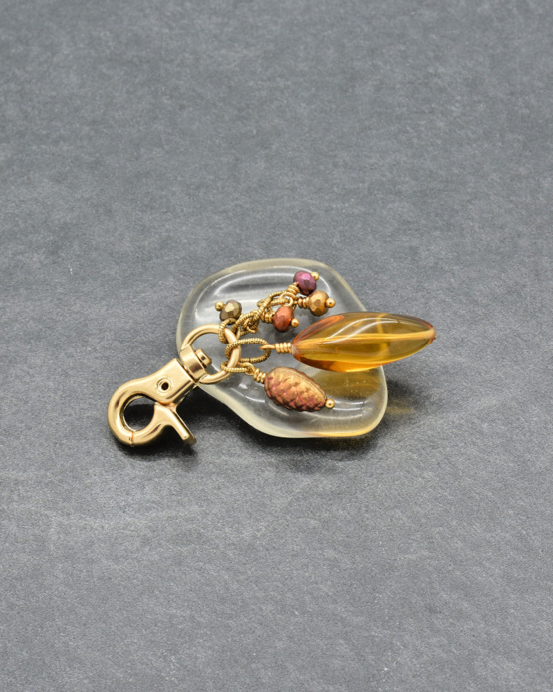 A decorative keychain or bag charm with a gold-tone lobster clasp, a clear teardrop-shaped acrylic holder, amber glass beads, a small brown pinecone-shaped bead, and tiny purple and orange accents, photographed on a dark gray textured background - made with love in Colorado by Ruby Acorn.