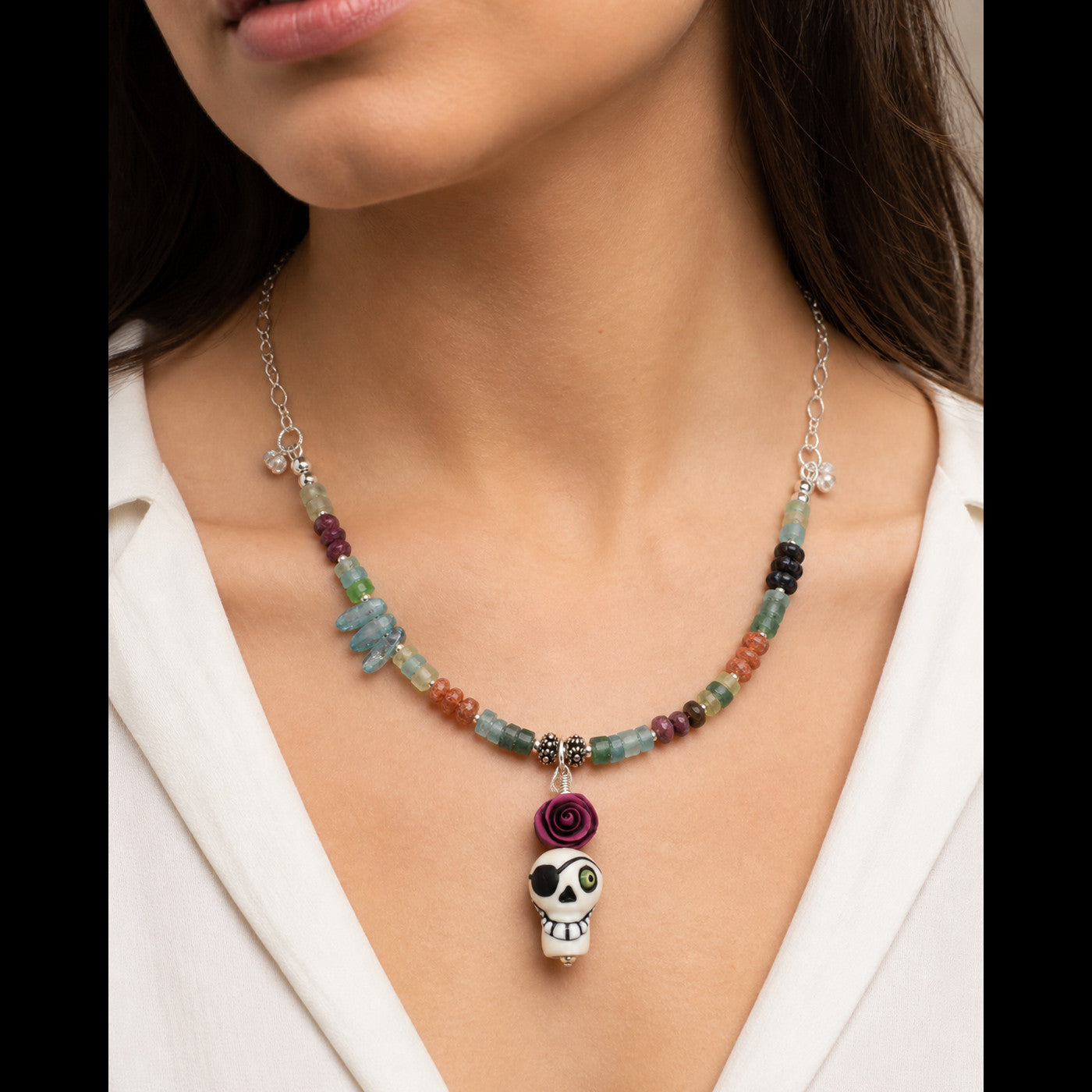 A close-up of a model wearing a beaded necklace with a sugar skull pendant. The necklace features a silver-tone chain and beads in various colors including green, blue, orange, purple, and black, with a white sugar skull charm and a purple rose accent.