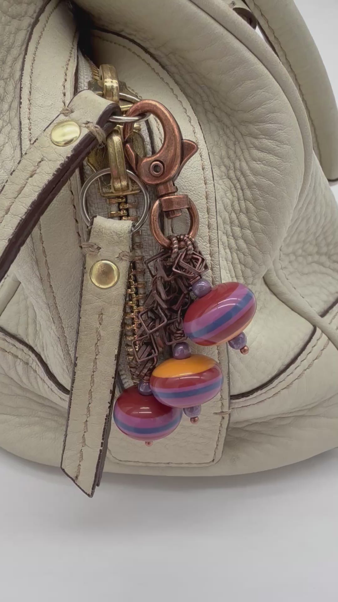 Video of close-up of a cream-colored leather purse featuring a decorative keychain charm with copper-toned hardware, purple beads, and striped vintage Lucite beads in pink, orange, and blue - made with love in Colorado by Ruby Acorn.