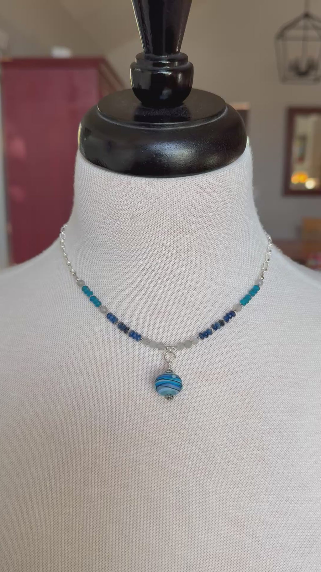 A video of a beaded necklace with blue and navy beads and a striped blue glass pendant, displayed on a white fabric mannequin bust.